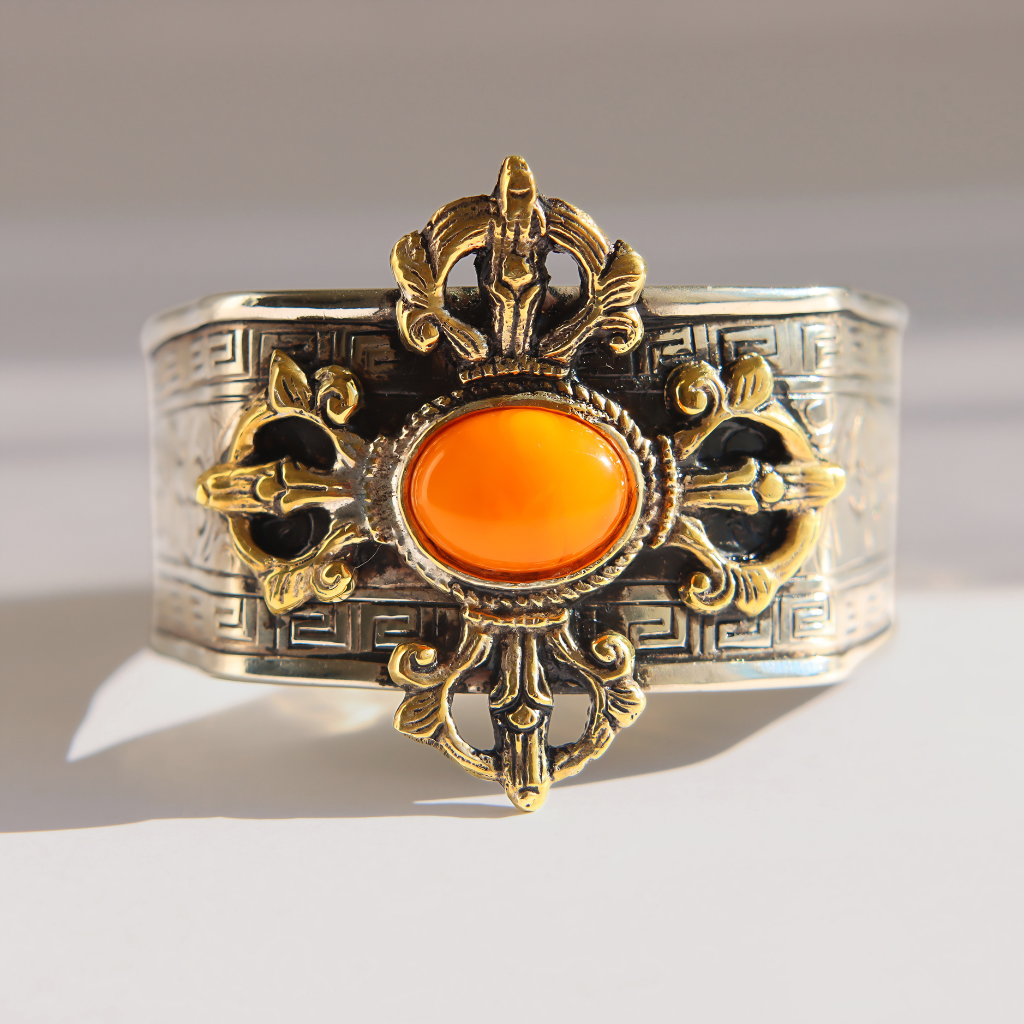 A wide silver-toned  amber cuff bracelet with ornate gold-toned filigree details and a central oval orange cabochon stone, photographed on a clean light gray background.