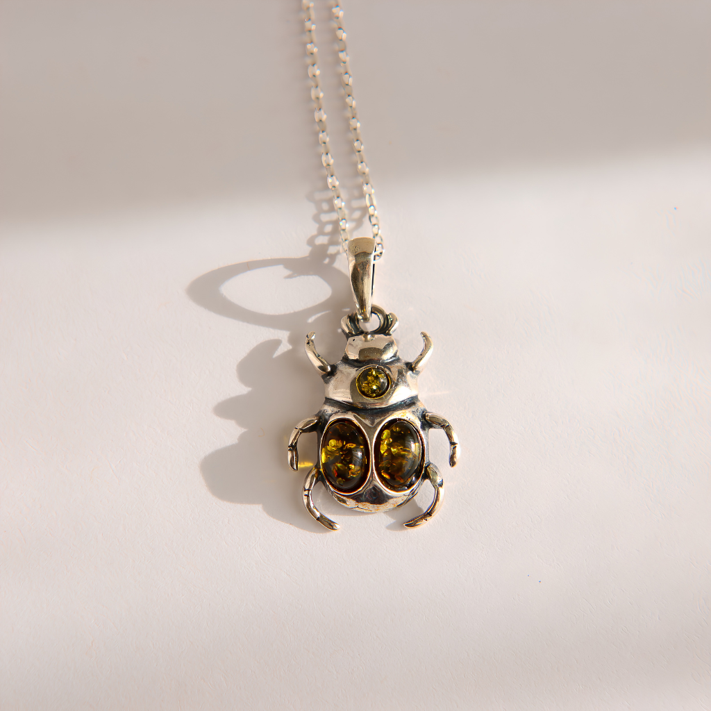 A silver amber beetle necklace with two oval amber-colored gemstones on the front and a smaller round amber stone above, hanging from a silver ball chain on a light background.