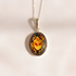 A silver chain amber scarab necklace with an oval pendant featuring a stylized scarab beetle design in yellow, orange, and red on a dark background, set against a plain off-white surface.