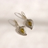 A pair of silver heart-shaped drop green amber earrings with intricate filigree cutouts and a central green marquise-cut stone, displayed on a light cream background.