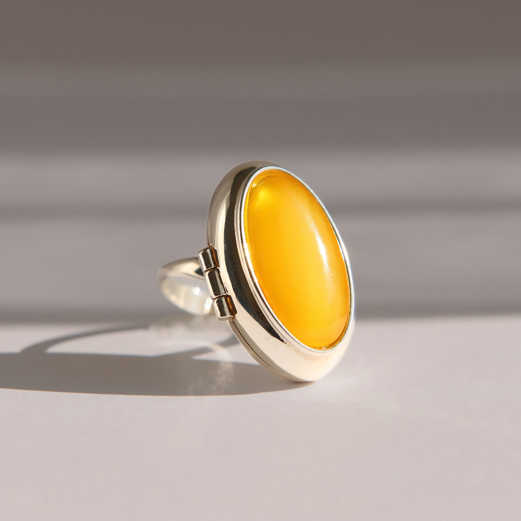 A sterling silver amber poison ring featuring a large, smooth, oval yellow stone, photographed on a neutral light gray background with soft shadows.