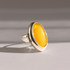 A sterling silver amber poison ring featuring a large, smooth, oval yellow stone, photographed on a neutral light gray background with soft shadows.