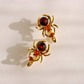A pair of gold spider-shaped earrings with dark red round gemstones and small orange-red accent crystals, photographed on a light cream background with soft shadows.