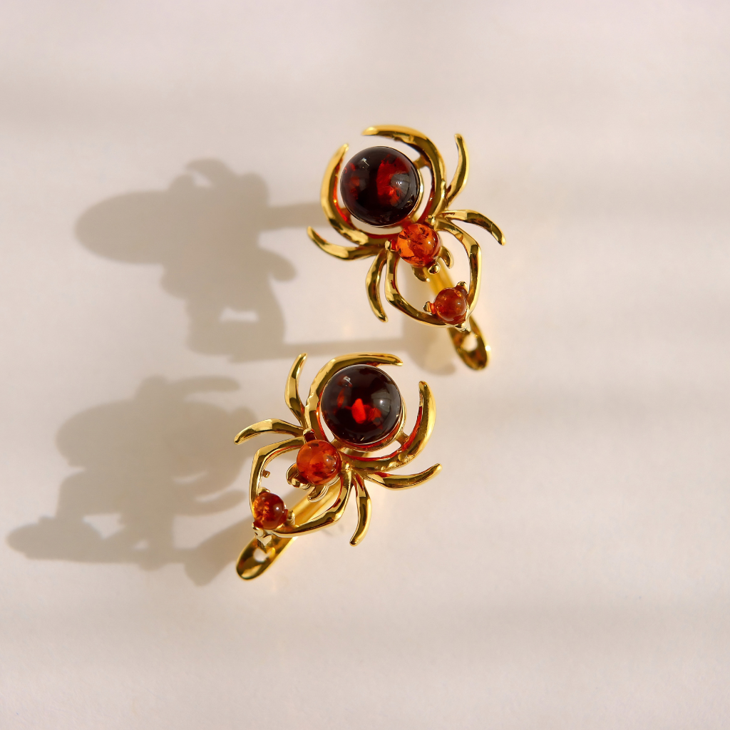 A pair of gold spider-shaped earrings with dark red round gemstones and small orange-red accent crystals, photographed on a light cream background with soft shadows.