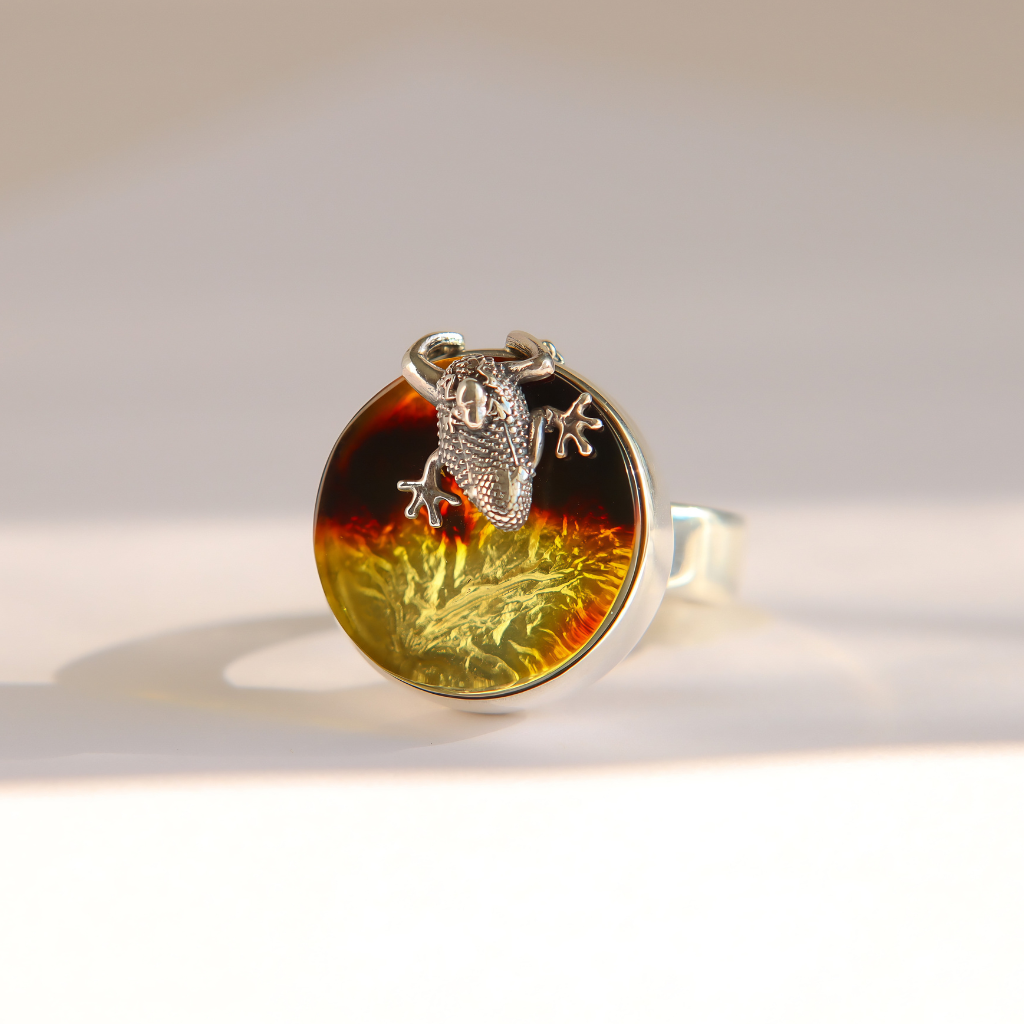 A silver amber ring featuring a round bezel-set amber cabochon with a gradient from dark brown to golden yellow, topped with a detailed silver frog figurine, photographed on a clean white surface with soft shadows.