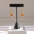 A pair of gold dangle spider amber earrings with orange gemstone centers, displayed on a black jewelry stand against a light gray background.
