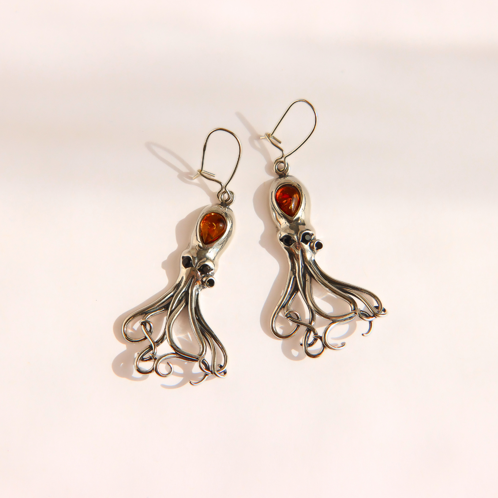 A pair of silver-toned amber octopus dangle earrings with polished amber-orange gemstones, displayed on a clean, light beige background.