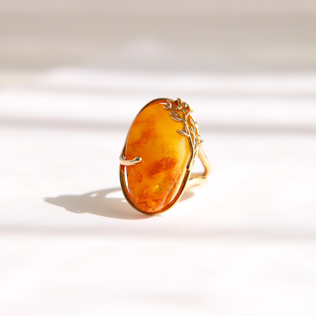 A gold amber ring featuring an organic-shaped amber cabochon with natural orange and yellow marbling, set against a bright, softly shadowed white background.
