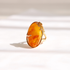 A gold amber ring featuring an organic-shaped amber cabochon with natural orange and yellow marbling, set against a bright, softly shadowed white background.