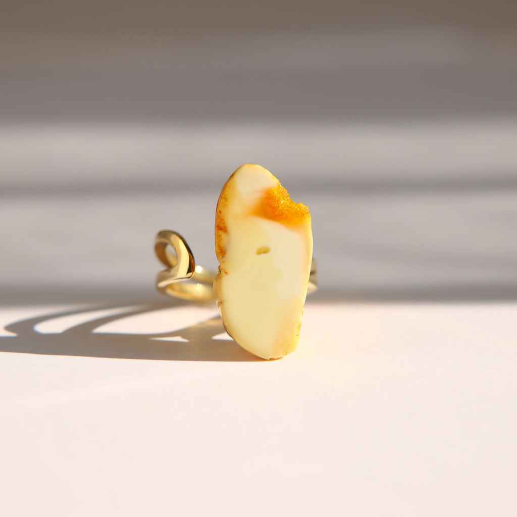 A gold white amber ring featuring a polished, irregularly shaped cream-colored stone with a vibrant orange-yellow fiery edge, photographed in dramatic direct sunlight on a light surface.