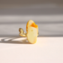 A gold white amber ring featuring a polished, irregularly shaped cream-colored stone with a vibrant orange-yellow fiery edge, photographed in dramatic direct sunlight on a light surface.