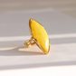 A gold butterscotch amber ring featuring a large, organic-shaped yellow stone, photographed on a light beige surface with soft diagonal shadows.