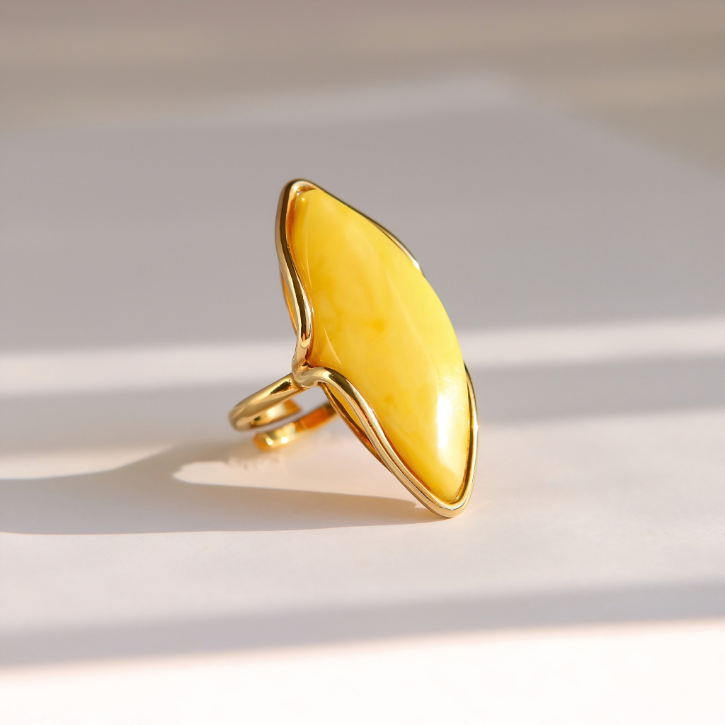 A gold butterscotch amber ring featuring a large, organic-shaped yellow stone, photographed on a light beige surface with soft diagonal shadows.