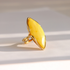 A gold butterscotch amber ring featuring a large, organic-shaped yellow stone, photographed on a light beige surface with soft diagonal shadows.