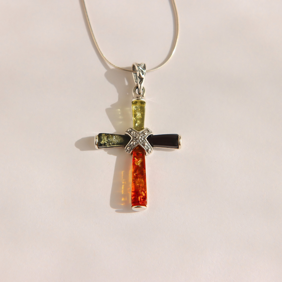 A silver amber cross pendant necklace featuring three vertical bars of amber in yellow-green, black, and orange-red, with a embellished center, suspended from a thin silver chain against a plain light background.