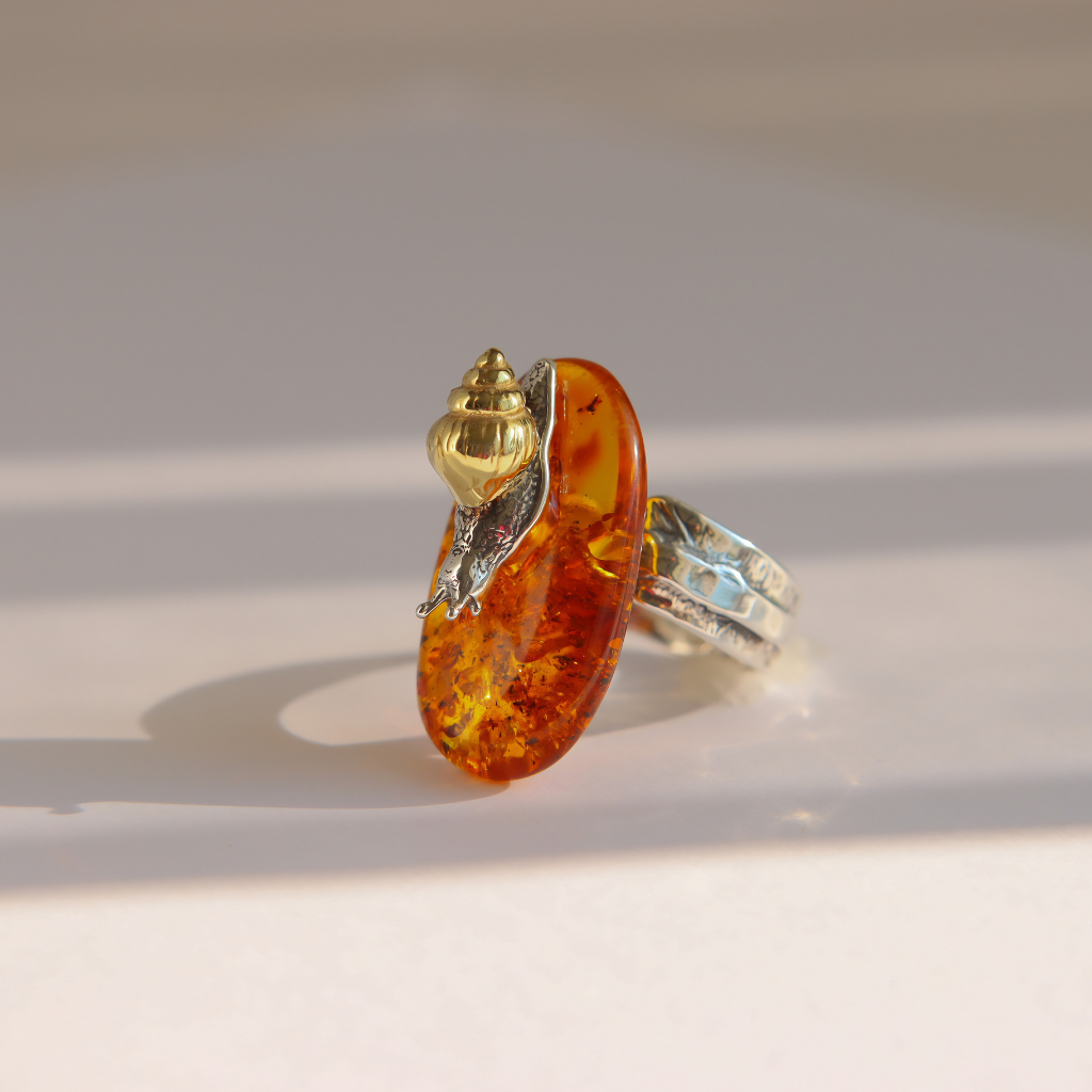 A unique amber ring featuring a large, polished amber-colored stone with inclusions, topped with a gold and silver snail set on a textured silver band, photographed on a light beige surface with soft shadows.