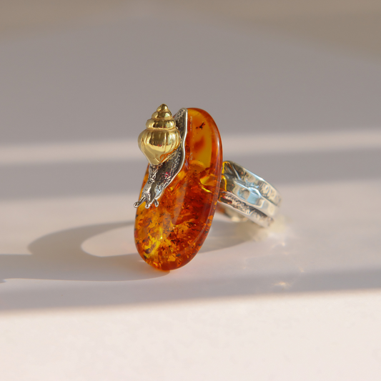 A unique amber ring featuring a large, polished amber-colored stone with inclusions, topped with a gold and silver snail set on a textured silver band, photographed on a light beige surface with soft shadows.