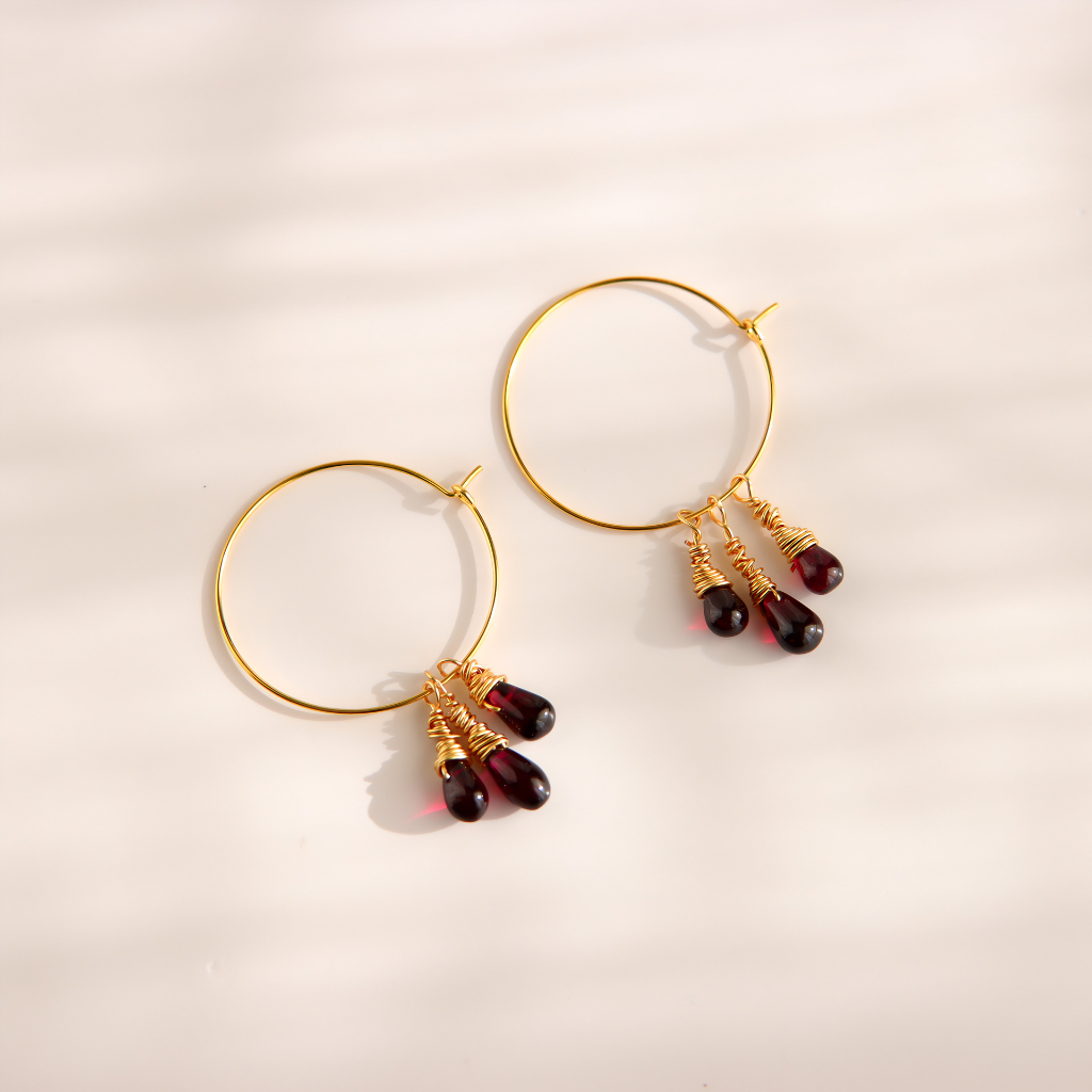 Gold hoop earrings with three dangling teardrop beads wrapped in gold wire, set against a warm off-white background with soft shadows.