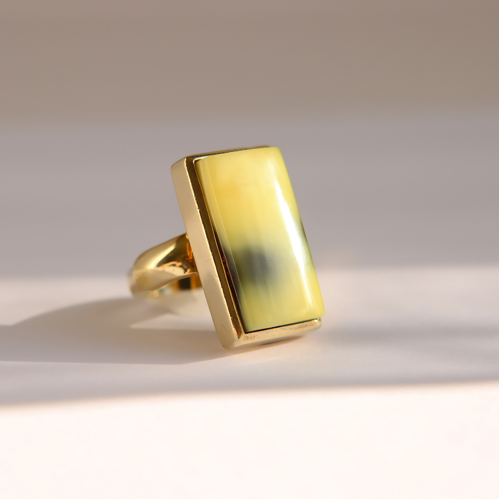 A gold Baltic amber ring with a large rectangular yellow-green stone, photographed on a light beige background with soft shadows.