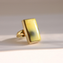 A gold Baltic amber ring with a large rectangular yellow-green stone, photographed on a light beige background with soft shadows.
