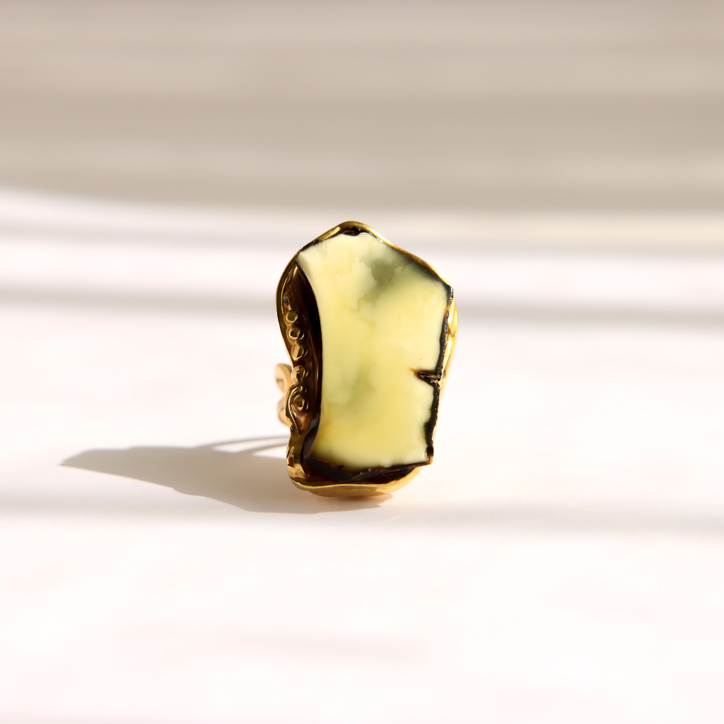 A gold amber statement ring with a large, irregularly shaped pale yellow-green stone, photographed on a clean white surface with soft shadows.
