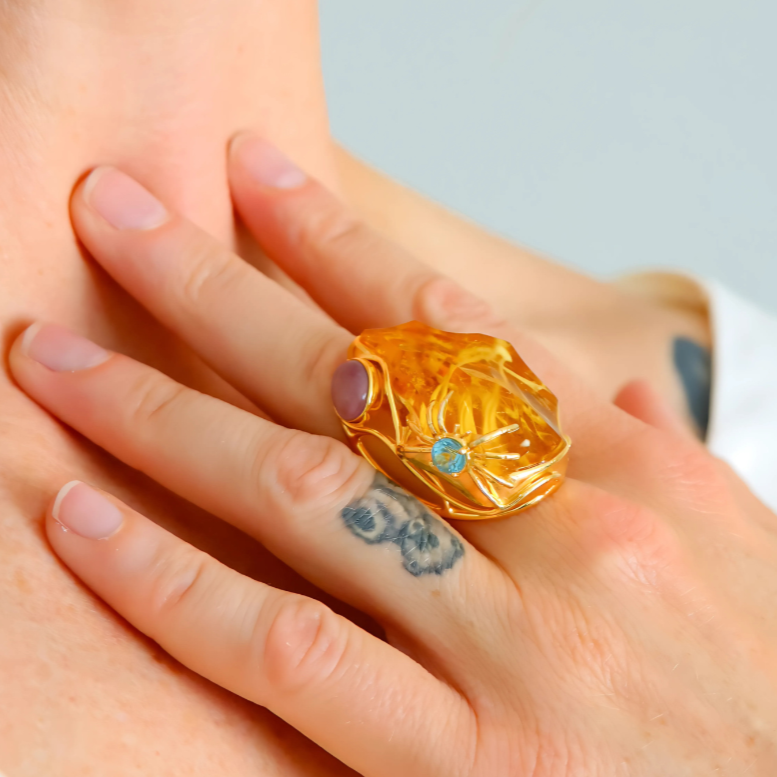 Gold cocktail ring with a blue gemstone on a person's finger against a neutral background