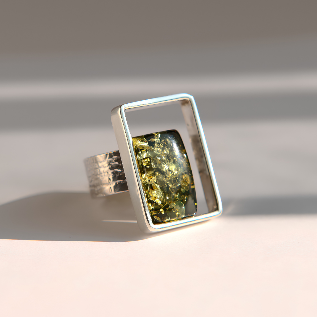 A silver amber ring with a textured band and a large rectangular frame holding a green marbled stone, photographed on a light beige surface with soft shadows.