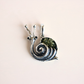 A silver amber snail pendant featuring a textured green shell and small black bead eyes, shown on a solid pale pink background.