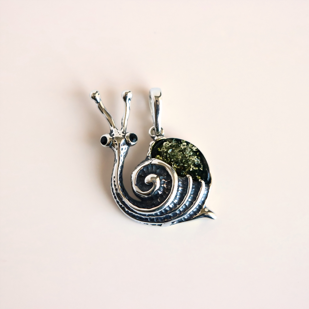 A silver amber snail pendant featuring a textured green shell and small black bead eyes, shown on a solid pale pink background.