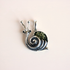 A silver amber snail pendant featuring a textured green shell and small black bead eyes, shown on a solid pale pink background.