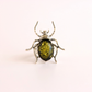 A beetle-shaped ring featuring a textured silver-toned metal body with legs and antennae, set with an oval green amber inlay on a plain light background.
