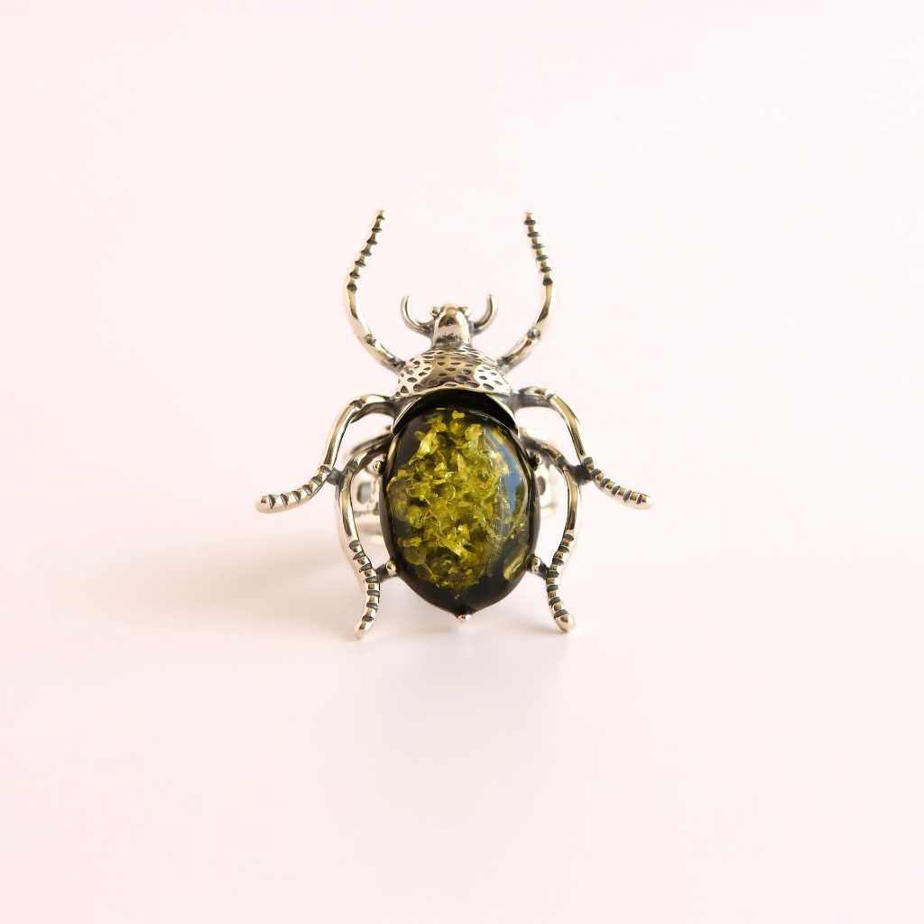 A beetle-shaped ring featuring a textured silver-toned metal body with legs and antennae, set with an oval green amber inlay on a plain light background.