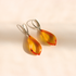 A pair of teardrop-shaped Baltic amber dangle earrings with silver leverback closures displayed on a warm beige background with soft shadows.
