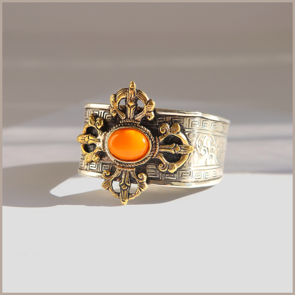An ornate silver-toned cuff amber bracelet with a central oval orange cabochon stone, surrounded by gold-toned filigree and Greek key detailing, photographed on a neutral light gray background.