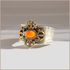 An ornate silver-toned cuff amber bracelet with a central oval orange cabochon stone, surrounded by gold-toned filigree and Greek key detailing, photographed on a neutral light gray background.