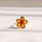 A silver amber flower ring with cognac amber-colored petals and a small red center stone, photographed on a light beige surface with soft shadows.