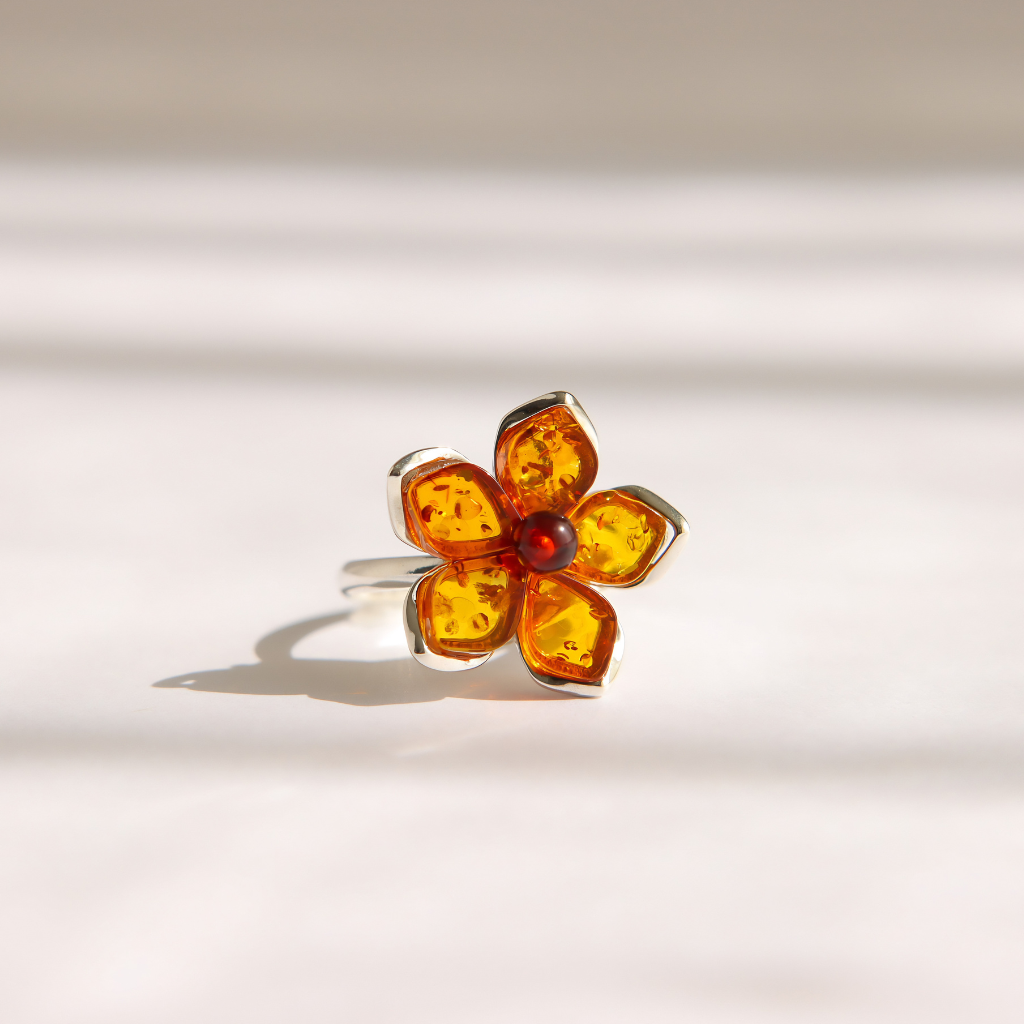 A silver amber flower ring with cognac amber-colored petals and a small red center stone, photographed on a light beige surface with soft shadows.