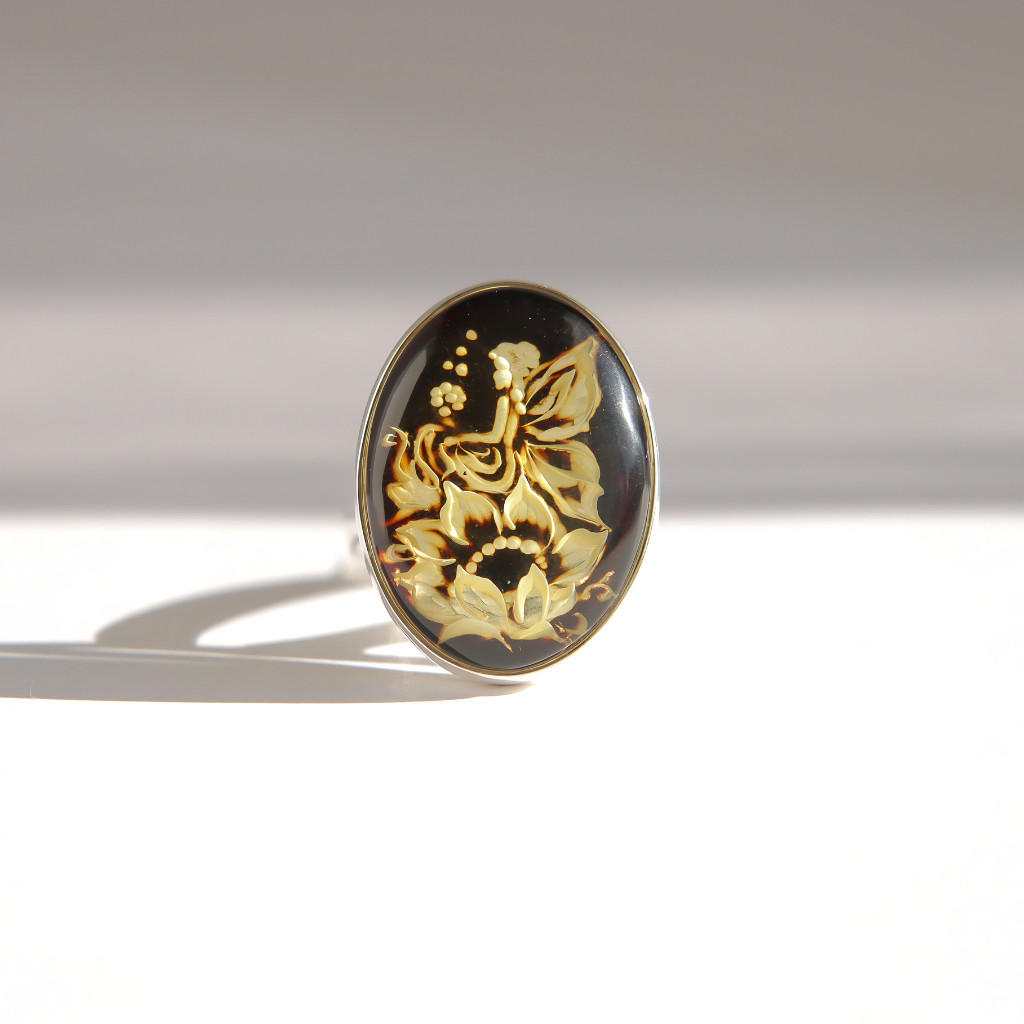 An oval amber cameo ring featuring a black amber background with carved floral and butterfly motifs, photographed on a clean white surface with a soft shadow.