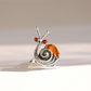 A snail-shaped ring made of sterling silver metal with a polished amber-orange shell and two small red eye stones, photographed on a clean light beige background.