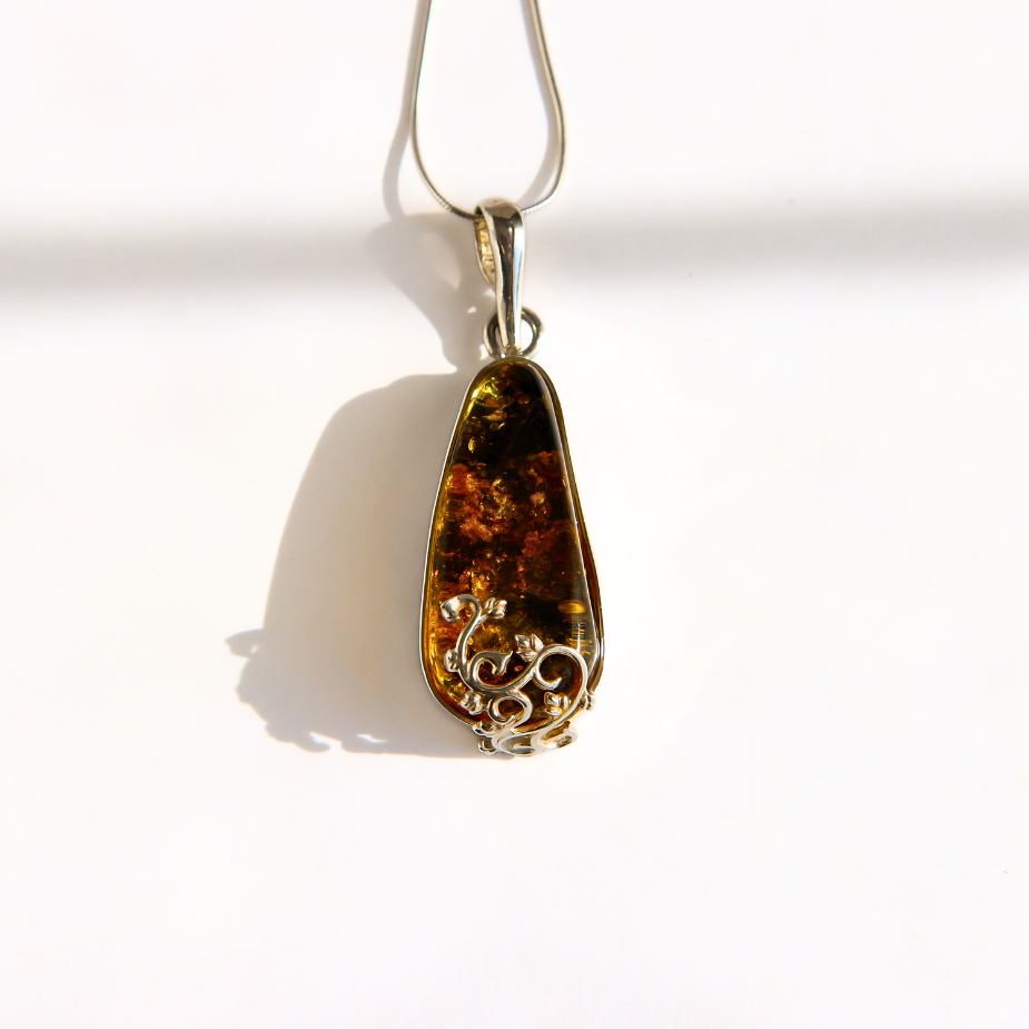 A teardrop-shaped green amber pendant with a silver-toned filigree-wrapped bottom, hanging from a silver chain against a bright white background.