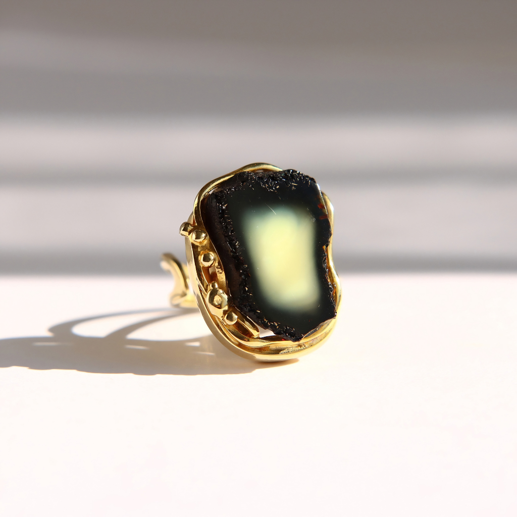 A gold amber ring featuring a large, irregular-shaped black amber stone with a yellow and white center, photographed on a white surface with soft shadows.