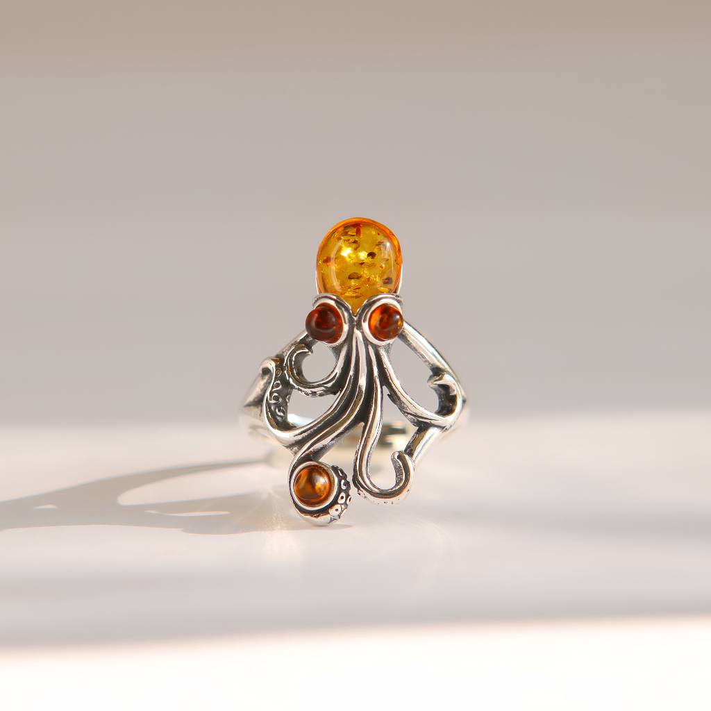 An ornate sterling silver amber octopus ring featuring a large round amber top stone and three smaller amber accent stones, photographed on a clean, light beige background.