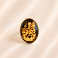 An oval amber cameo ring featuring a black background with a detailed gold illustration of a fairy and flowers, set in a gold-toned metal bezel, displayed on a soft beige satin fabric.