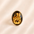 An oval amber cameo ring featuring a black background with a detailed gold illustration of a fairy and flowers, set in a gold-toned metal bezel, displayed on a soft beige satin fabric.