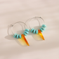 A pair of silver and turquoise hoop earrings with light blue beads and amber-orange drop charms, displayed on a warm beige background with soft shadows.