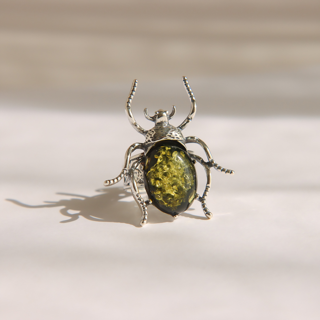 A silver beetle ring with a textured, metallic body and a prominent oval green amber gemstone on the abdomen, set against a warm beige background with a soft shadow.