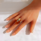 A close-up of a hand wearing a gold amber ring with a polished amber-orange oval stone, set against a bright white background.