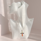 A silver amber cross pendant necklace with colorful enamel details is displayed on a white mannequin bust against a soft pink background.