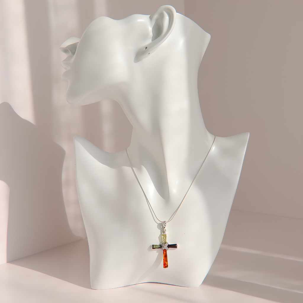 A silver amber cross pendant necklace with colorful enamel details is displayed on a white mannequin bust against a soft pink background.