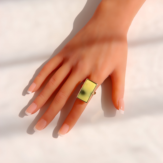 A gold-toned rectangular Baltic amber ring with a green reflective center, worn on a hand with a French manicure against a white background with soft shadows.